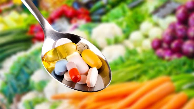 Spoon filled with various dietary supplement capsules and tablets with fresh vegetables blurred in the background