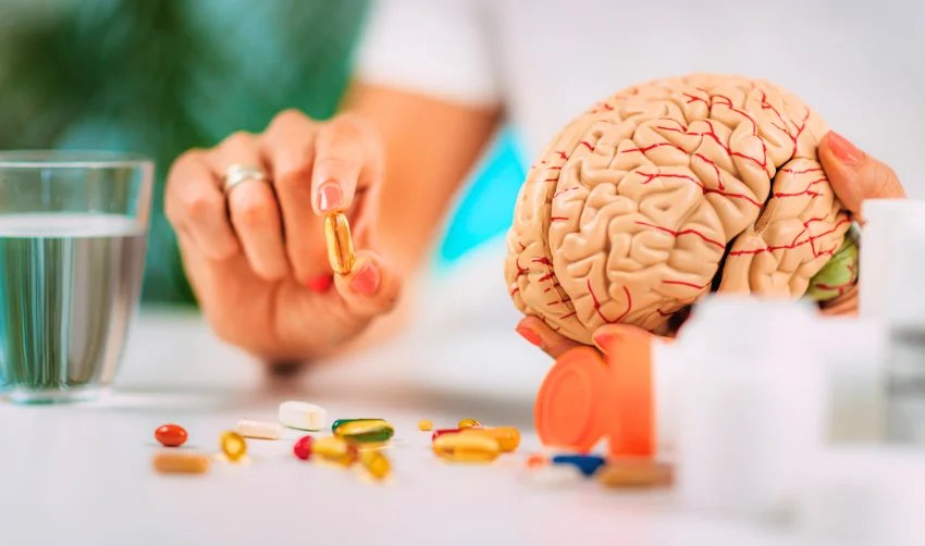 Hands holding a brain model and a softgel with supplements nearby
